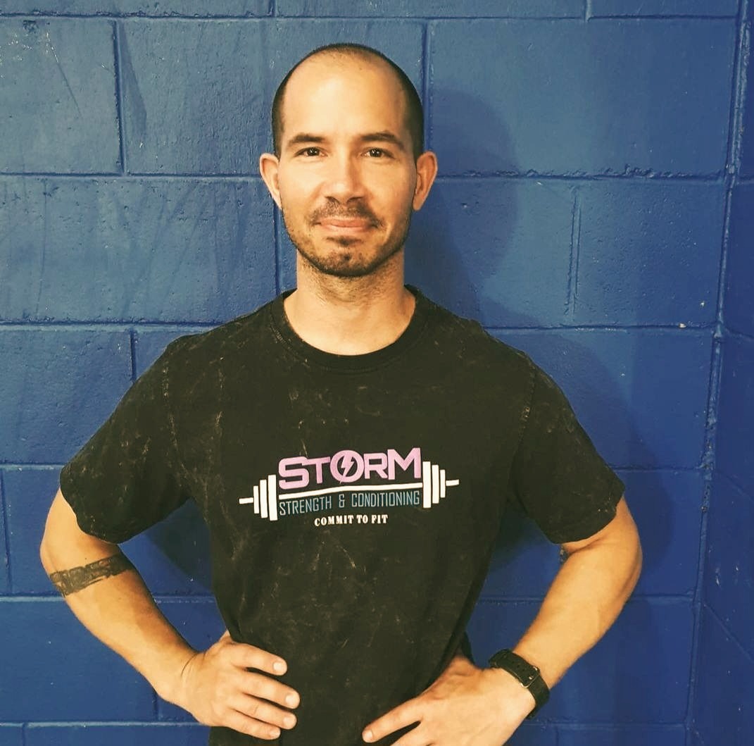 Coach Todd standing in a Storm shirt in front of the blue gym wall
